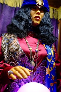 A mannequin dressed in an elaborate fortune teller costume, featuring a dark, voluminous wig and a deep blue turban adorned with a large jewel. The outfit is richly patterned with swirling designs and star motifs, primarily in purple, blue, and gold hues. The mannequin is posed with one hand hovering over a glowing crystal ball, adding a mystical ambiance.