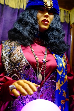 A mannequin dressed in an elaborate fortune teller costume, featuring a dark, voluminous wig and a deep blue turban adorned with a large jewel. The outfit is richly patterned with swirling designs and star motifs, primarily in purple, blue, and gold hues. The mannequin is posed with one hand hovering over a glowing crystal ball, adding a mystical ambiance.