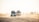 A group of off-road vehicles driving over sandy dunes under bright daylight. Several people are riding in and on top of the vehicles, possibly participating in a safari or desert tour. The background shows a hazy horizon with scattered silhouettes of more vehicles and people.