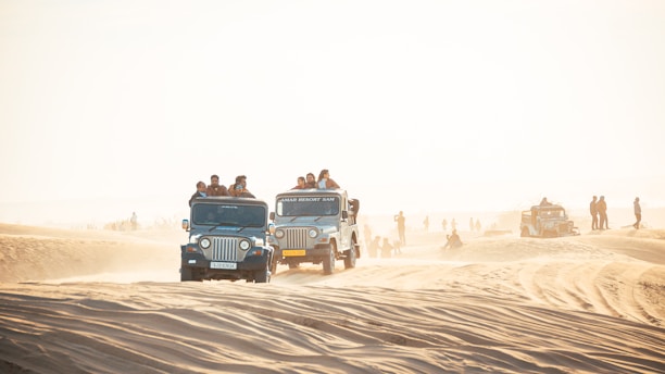 A group of off-road vehicles driving over sandy dunes under bright daylight. Several people are riding in and on top of the vehicles, possibly participating in a safari or desert tour. The background shows a hazy horizon with scattered silhouettes of more vehicles and people.