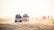 A group of off-road vehicles driving over sandy dunes under bright daylight. Several people are riding in and on top of the vehicles, possibly participating in a safari or desert tour. The background shows a hazy horizon with scattered silhouettes of more vehicles and people.