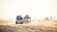 A group of off-road vehicles driving over sandy dunes under bright daylight. Several people are riding in and on top of the vehicles, possibly participating in a safari or desert tour. The background shows a hazy horizon with scattered silhouettes of more vehicles and people.