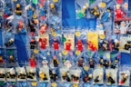 A collection of various LEGO minifigures individually packed in small plastic bags, displayed on a flat blue surface. Each bag contains a different minifigure, some labeled with handwritten tags and prices. The minifigures are in different costumes and roles, including racers, firefighters, and other characters.