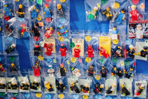 A collection of various LEGO minifigures individually packed in small plastic bags, displayed on a flat blue surface. Each bag contains a different minifigure, some labeled with handwritten tags and prices. The minifigures are in different costumes and roles, including racers, firefighters, and other characters.