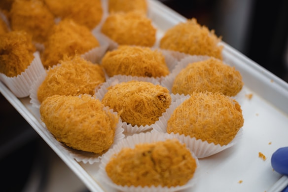 A tray filled with golden-brown, crispy fried pastries, each placed in a white paper cup. The pastries have a flaky, spiral texture, suggesting a light and airy consistency.