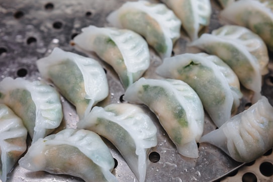 A group of translucent dumplings is arranged on a perforated metal steamer tray. The dumplings have a delicate, translucent skin and appear to be filled with a mixture containing green fillings, likely vegetables or herbs. The surface reflects a fresh, slightly shiny appearance.