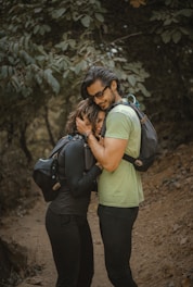 A couple embraces warmly on a forested trail. They are wearing hiking gear, including backpacks. The path is surrounded by lush green foliage, creating a serene and intimate atmosphere.