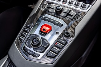 A sleek car interior showcasing a high-tech central control panel featuring a mix of tactile buttons and switches, surrounded by carbon fiber trim. Prominent buttons include options for navigation, media, and driving modes with a bold red start button.