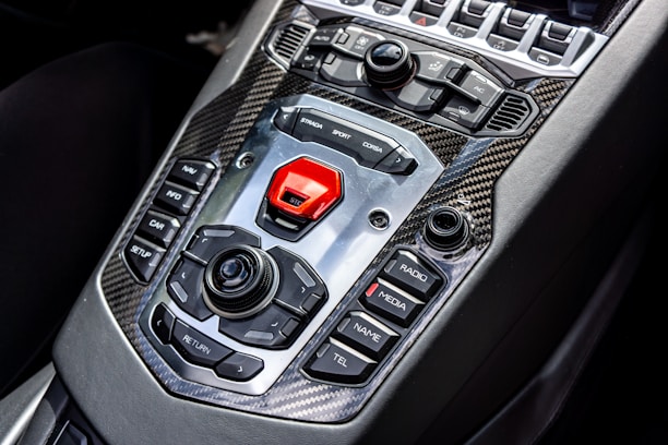 A sleek car interior showcasing a high-tech central control panel featuring a mix of tactile buttons and switches, surrounded by carbon fiber trim. Prominent buttons include options for navigation, media, and driving modes with a bold red start button.