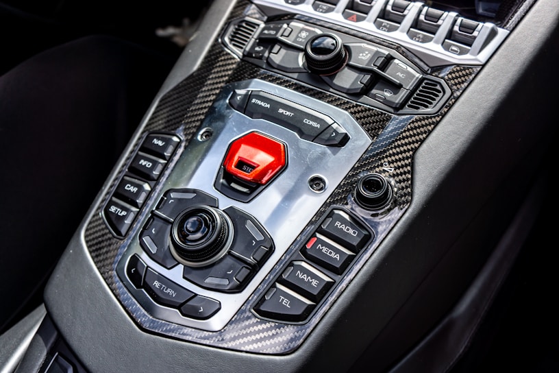 A sleek car interior showcasing a high-tech central control panel featuring a mix of tactile buttons and switches, surrounded by carbon fiber trim. Prominent buttons include options for navigation, media, and driving modes with a bold red start button.
