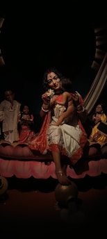 The image depicts a group of colorful, traditional statues. The central figure, a woman dressed in ornate garments, holds a child. Her attire is red and white with intricate patterns. There are several other figures around her, each adorned in vibrant costumes. The scene conveys a cultural or religious significance with an emphasis on heritage and tradition.