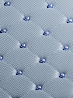A detailed pattern of tufted fabric with smooth, rounded upholstery buttons. The fabric has a textured, mesh-like appearance and is neatly stitched, creating a grid of soft, cushioned sections.
