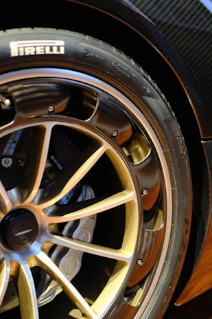 A close-up view of a car wheel featuring a gold alloy rim with a detailed spoke design. The Pirelli tire branding is visible on the outer edge of the tire. The brake caliper can be seen behind the rim.