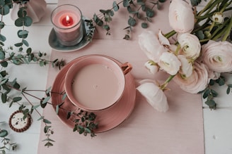 A serene scene featuring a cup of tea on a pink saucer surrounded by white flowers and lush green eucalyptus leaves. A lit candle in a glass holder sits nearby, creating a cozy atmosphere. A single cookie is placed to the side, enhancing the inviting setting.
