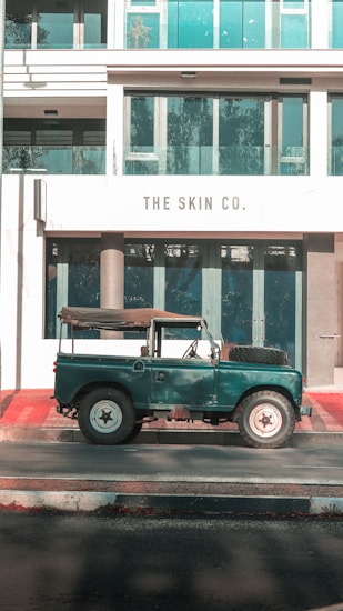 A vintage green off-road vehicle is parked on a street in front of a modern building with large glass windows and a sign reading 'The Skin Co.'. The building features clean architectural lines, and the sunlight casts soft shadows on the scene.