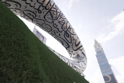 A modern, curvilinear architectural structure with Arabic calligraphy stands prominently against a bright sky. A tall, angular building with a unique design is visible in the background. The foreground features green, well-maintained grass.