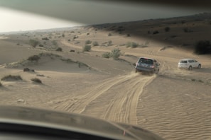 A desert landscape with sand dunes featuring two off-road vehicles driving on the tracks. Sparse vegetation is scattered across the sandy terrain, and tire tracks are visible behind the vehicles.