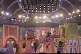 A vibrant stage performance featuring a group of people in colorful, traditional attire. The stage is decorated with ornate, Indian-style architecture and bright lighting, creating a festive atmosphere. The performers are dynamically posed, some descending stairs while others appear mid-action on a multi-level set.
