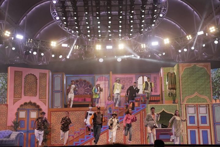 A vibrant stage performance featuring a group of people in colorful, traditional attire. The stage is decorated with ornate, Indian-style architecture and bright lighting, creating a festive atmosphere. The performers are dynamically posed, some descending stairs while others appear mid-action on a multi-level set.