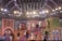 A vibrant stage performance featuring a group of people in colorful, traditional attire. The stage is decorated with ornate, Indian-style architecture and bright lighting, creating a festive atmosphere. The performers are dynamically posed, some descending stairs while others appear mid-action on a multi-level set.
