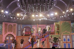 A vibrant stage performance featuring a group of people in colorful, traditional attire. The stage is decorated with ornate, Indian-style architecture and bright lighting, creating a festive atmosphere. The performers are dynamically posed, some descending stairs while others appear mid-action on a multi-level set.
