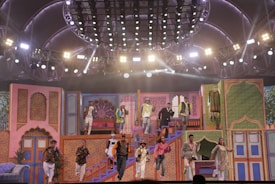 A vibrant stage performance featuring a group of people in colorful, traditional attire. The stage is decorated with ornate, Indian-style architecture and bright lighting, creating a festive atmosphere. The performers are dynamically posed, some descending stairs while others appear mid-action on a multi-level set.