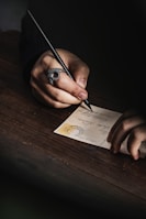 A person is writing on a postcard using a black pen. The hand features a large, ornate ring, and the postcard has a visible stamp and handwriting on it. The atmosphere is intimate, with a focus on the act of writing.