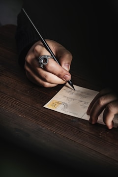 A person is writing on a postcard using a black pen. The hand features a large, ornate ring, and the postcard has a visible stamp and handwriting on it. The atmosphere is intimate, with a focus on the act of writing.