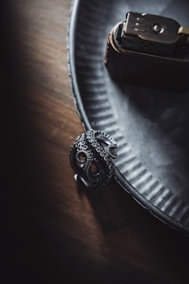 A metal octopus-shaped ring rests on a textured, gray plate adjacent to a metallic lighter. The lighting casts soft shadows, enhancing the intricate details of the ring and the textures of the surfaces.