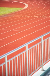 A curved running track with several parallel lanes is visible, featuring vibrant red surface and white lane markings. A railing is in the foreground, indicating a boundary or barrier. A patch of green grass is seen beside the track.