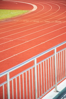 A curved running track with several parallel lanes is visible, featuring vibrant red surface and white lane markings. A railing is in the foreground, indicating a boundary or barrier. A patch of green grass is seen beside the track.