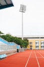 A sports stadium with a red running track, blue seating area, and a large floodlight. The background features a yellow building with blue and white accents, surrounded by green trees. The atmosphere is clean and empty, with clear skies above.