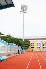 A sports stadium with a red running track, blue seating area, and a large floodlight. The background features a yellow building with blue and white accents, surrounded by green trees. The atmosphere is clean and empty, with clear skies above.