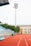 A sports stadium with a red running track, blue seating area, and a large floodlight. The background features a yellow building with blue and white accents, surrounded by green trees. The atmosphere is clean and empty, with clear skies above.