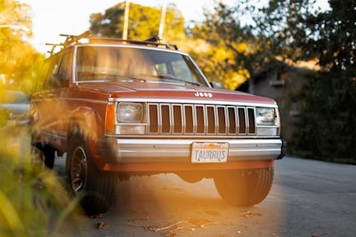 A vintage red Jeep with a front grill and a California license plate reading 'TaurRUS' is parked on the side of a street. The car is surrounded by greenery and bathed in warm sunlight, creating a soft, golden lens flare effect.