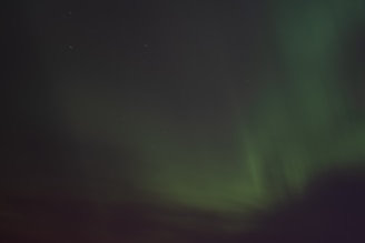 A night sky illuminated by sweeping bands of green light, consistent with the aurora borealis. Stars are faintly visible in the background, adding to the celestial atmosphere.