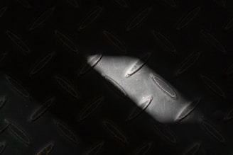 A section of dark metal with oval-shaped patterns and a narrow beam of light casting a shadow across the surface. The texture appears rough and industrial.