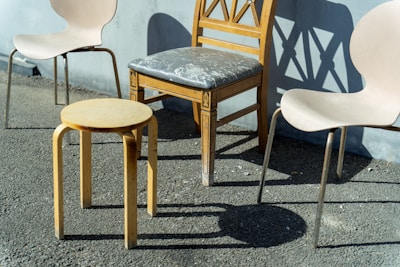 A collection of chairs arranged on a rough paved surface, consisting of a modern wooden stool, two light-colored plastic chairs with metallic legs, and a vintage wooden chair with a carved backrest and a worn seat cushion. The scene is bathed in natural sunlight, casting strong shadows on the surface.