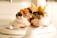 Two meringue desserts are presented on a white pedestal cake stand. Each is topped with a variety of toppings including whipped cream, crushed nuts, blueberries, a small orange fruit with husk, and pieces of dried orange peel.