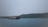 A serene waterbody is in the foreground, with a large traditional houseboat floating on it. In the background, a densely wooded shoreline with gentle hills creates a calm and peaceful atmosphere. The sky is overcast, adding a muted touch to the scene.