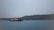 A serene waterbody is in the foreground, with a large traditional houseboat floating on it. In the background, a densely wooded shoreline with gentle hills creates a calm and peaceful atmosphere. The sky is overcast, adding a muted touch to the scene.