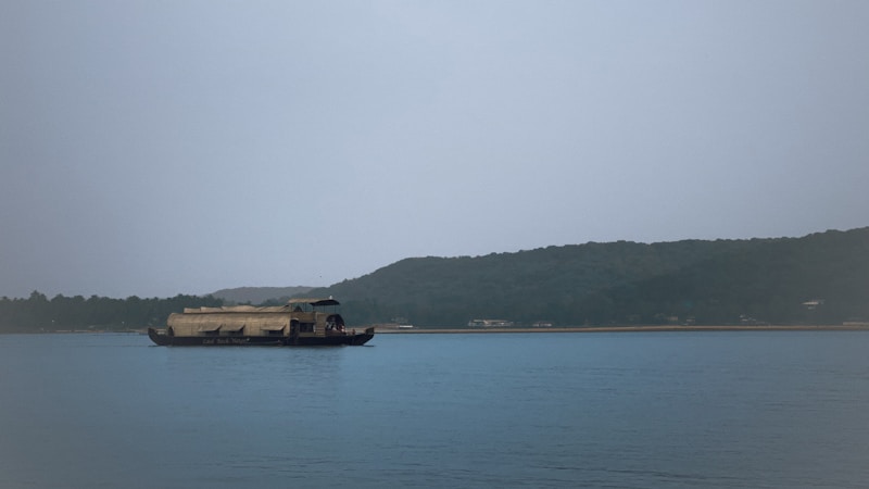A serene waterbody is in the foreground, with a large traditional houseboat floating on it. In the background, a densely wooded shoreline with gentle hills creates a calm and peaceful atmosphere. The sky is overcast, adding a muted touch to the scene.