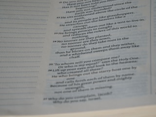 A page from a book containing biblical text with numbered verses. The right side of the page features text in a two-column format, while the left side shows faint dotted lines without text. The background is an off-white color with the text primarily in black, and there is a slight shadow over parts of the page.