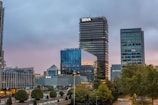 A cityscape featuring modern office buildings with reflective glass facades in a business district. In the foreground, well-maintained greenery and small trees are visible, highlighting urban landscaping. The sky is overcast, suggesting a transition from day to evening, casting a subdued light on the buildings.