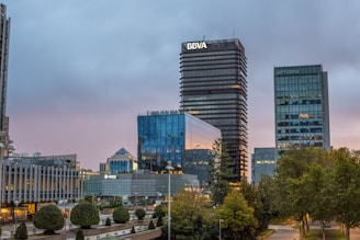 A cityscape featuring modern office buildings with reflective glass facades in a business district. In the foreground, well-maintained greenery and small trees are visible, highlighting urban landscaping. The sky is overcast, suggesting a transition from day to evening, casting a subdued light on the buildings.