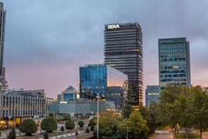A cityscape featuring modern office buildings with reflective glass facades in a business district. In the foreground, well-maintained greenery and small trees are visible, highlighting urban landscaping. The sky is overcast, suggesting a transition from day to evening, casting a subdued light on the buildings.