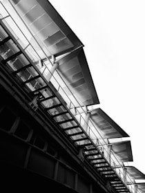 A black and white photograph of an architectural structure featuring an angled view of a bridge or overpass with multiple metallic beams and panels. The design includes columns supporting the structure and railings along the edges, creating strong lines and a modern, industrial aesthetic against a bright sky.