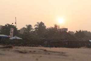 The image features a beachside scene at sunset with palm trees and a thatched-roof structure, possibly a small shop or cafe. The sun is setting in the distance, casting a warm glow over the area. There are umbrellas, chairs, and decorations such as strings of red and white lanterns along the beach.