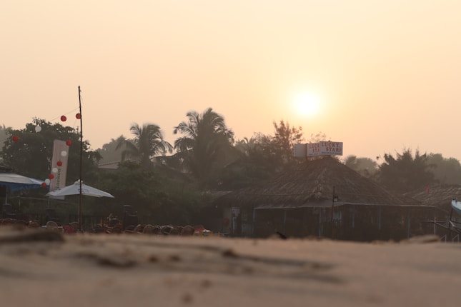 The image features a beachside scene at sunset with palm trees and a thatched-roof structure, possibly a small shop or cafe. The sun is setting in the distance, casting a warm glow over the area. There are umbrellas, chairs, and decorations such as strings of red and white lanterns along the beach.