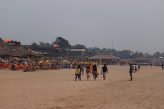 A sandy beach with several people walking along the shore, some in groups and others alone. On the left side, there are brightly colored beach chairs and huts with signage indicating restaurants or bars. The sky is overcast, adding a muted tone to the scene. In the background, trees and additional structures are visible.